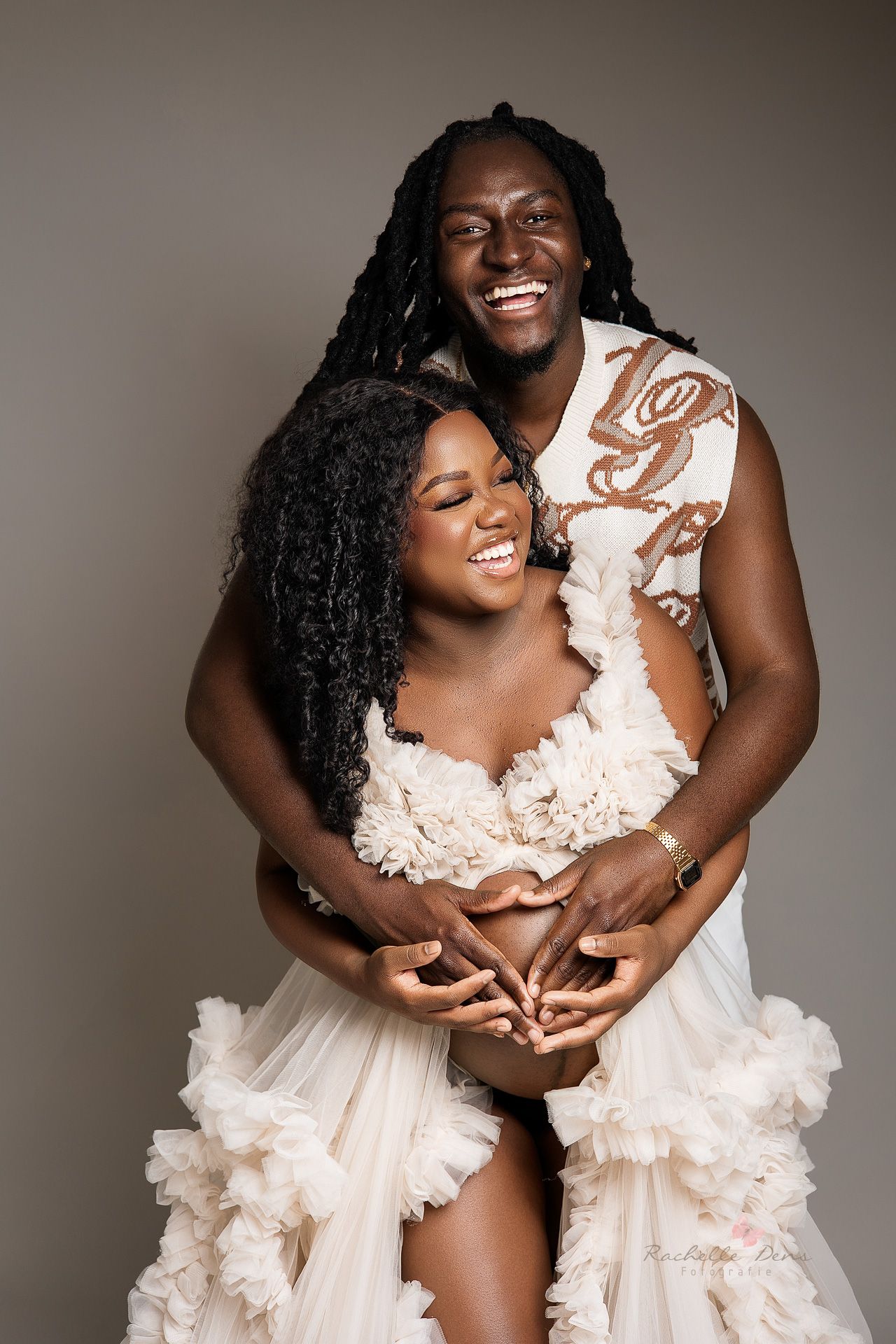 A romantic couple in white outfits embrace while smiling against a neutral backdrop in an intimate portrait session.
