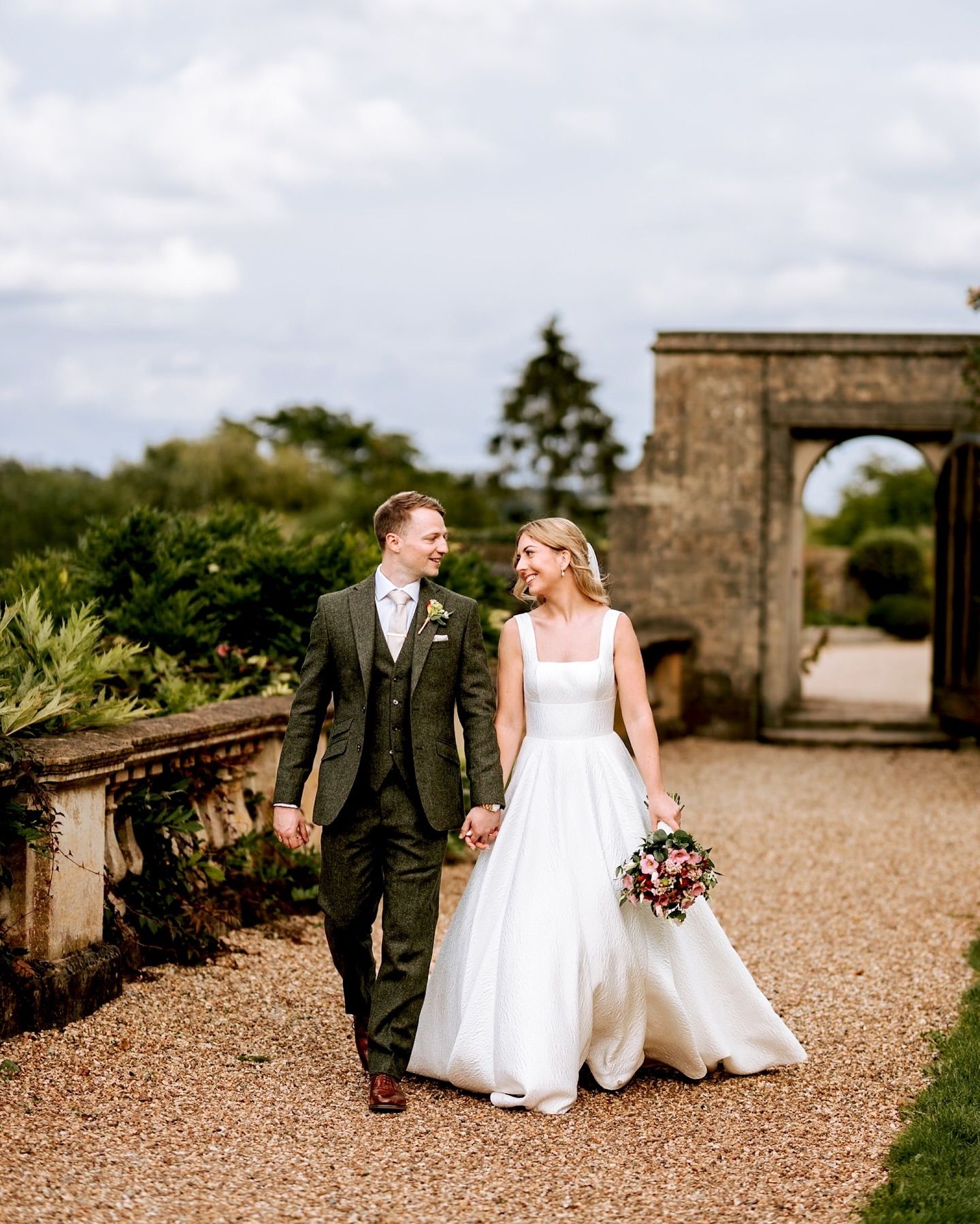 A couple walks hand in hand on a gravel path leading to a stone archway in a romantic garden setting.