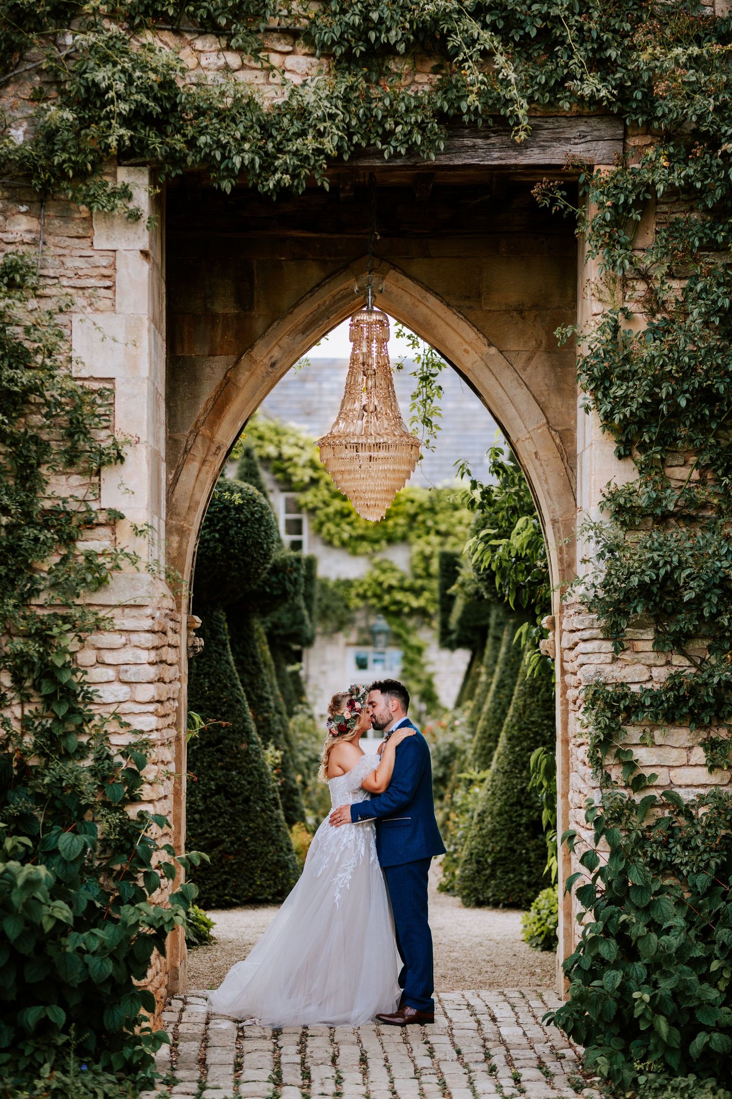Stone archway frames romantic wedding portrait with chandelier visible through garden path.
