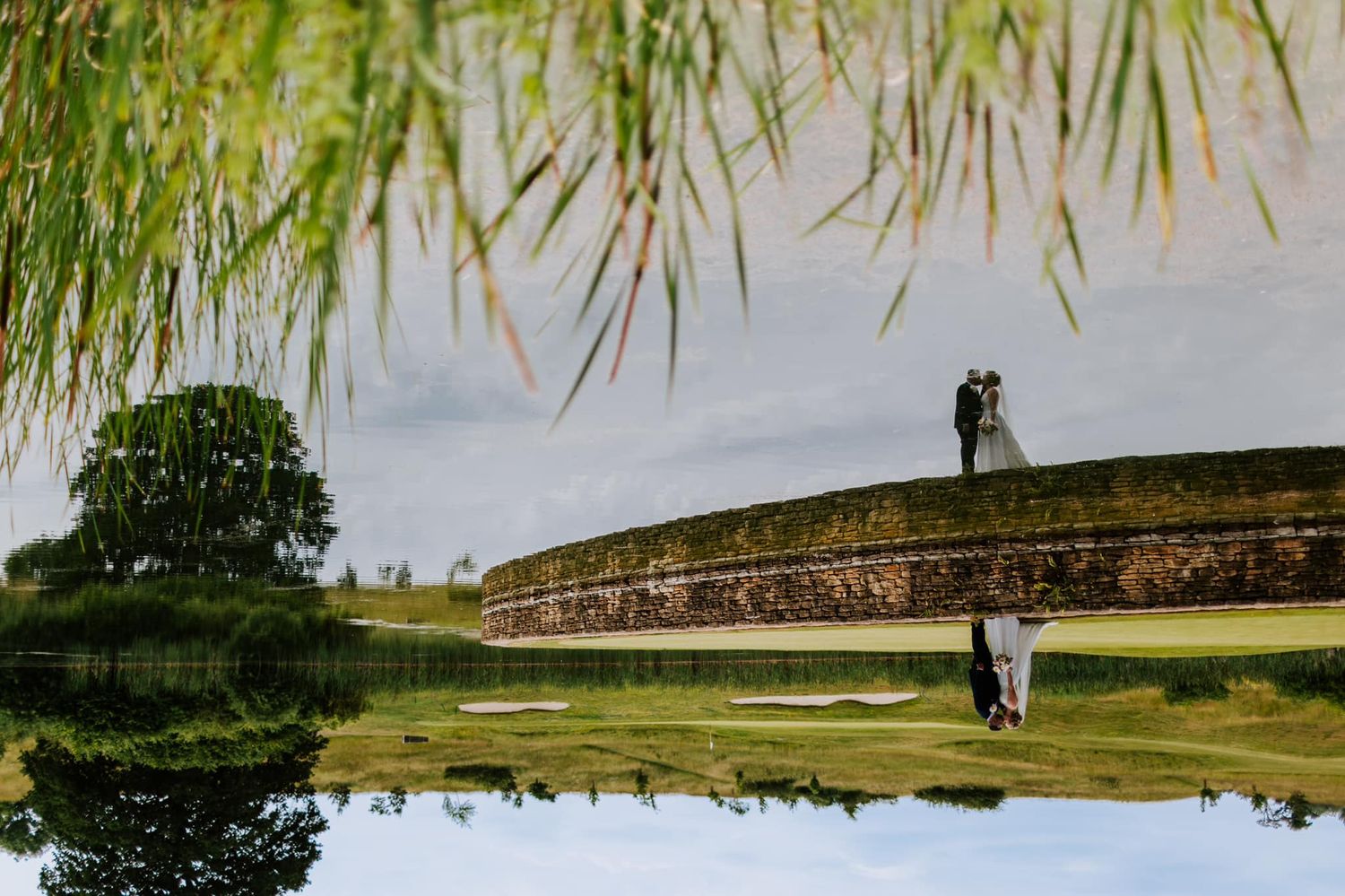 Artistic reflection in pond shows couple on stone bridge with greenery.