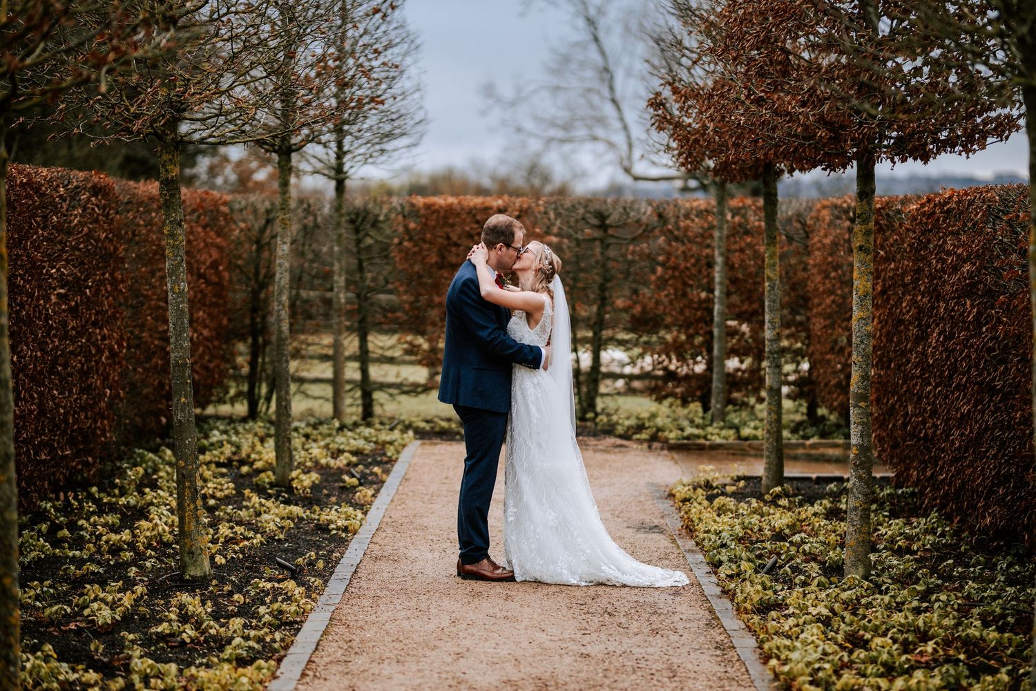 Wedding couple embraces on garden path lined with manicured hedges in autumn.