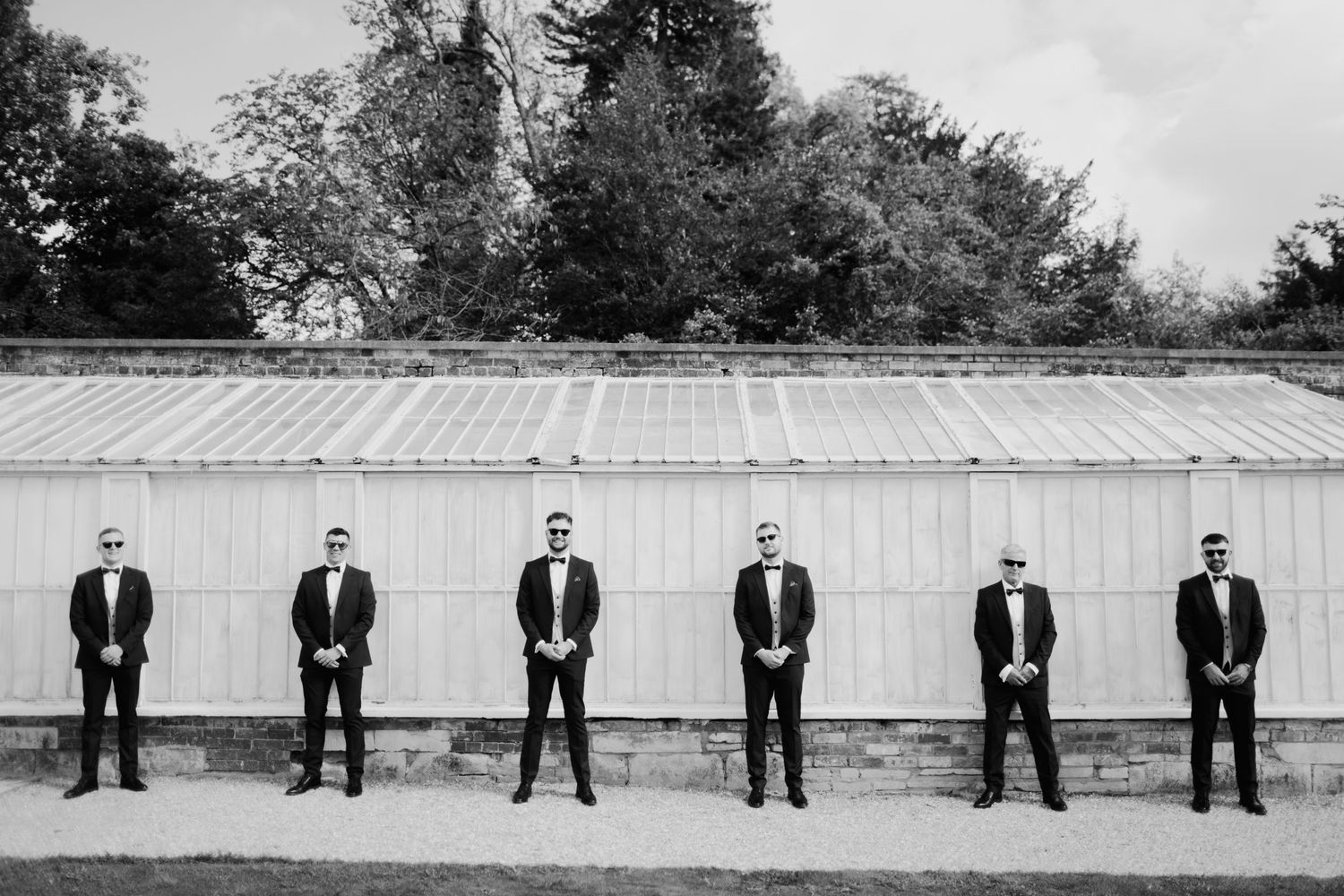 Groomsmen lined up in black tuxedos against white greenhouse background.