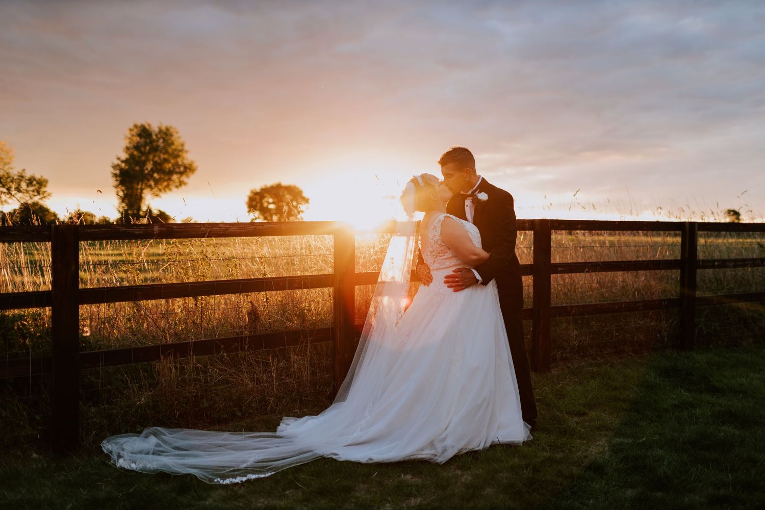 A romantic sunset silhouette of newlyweds by a wooden fence in the countryside.