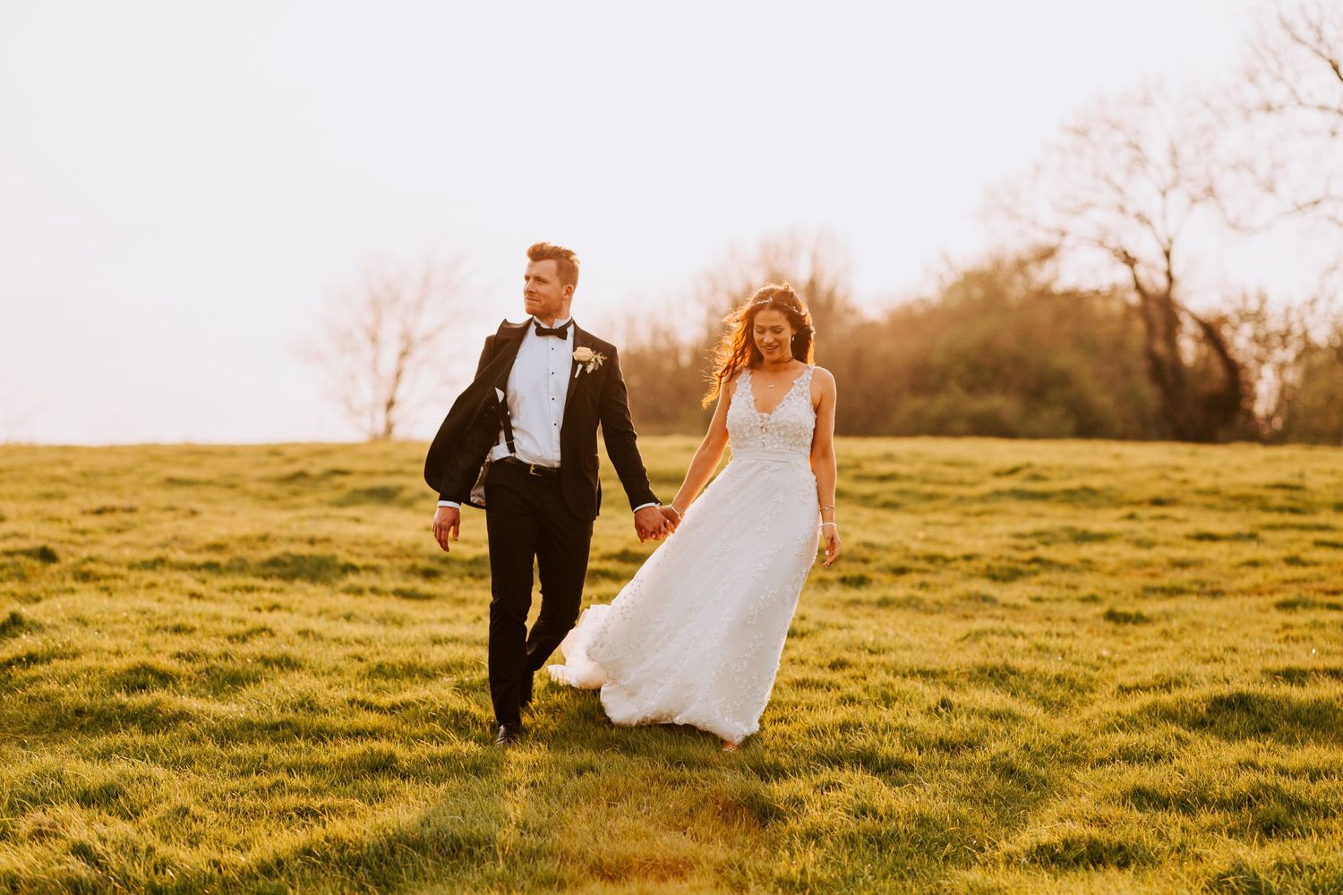 A couple in formal attire runs joyfully through a sunlit field.