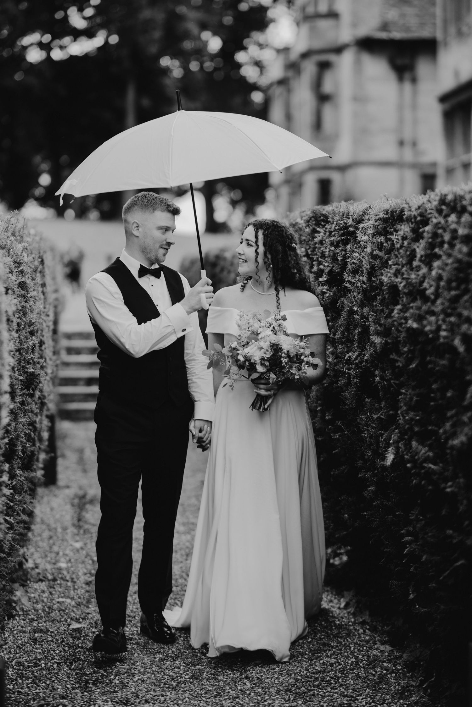 A romantic black and white wedding photo of a couple under an umbrella surrounded by garden hedges.