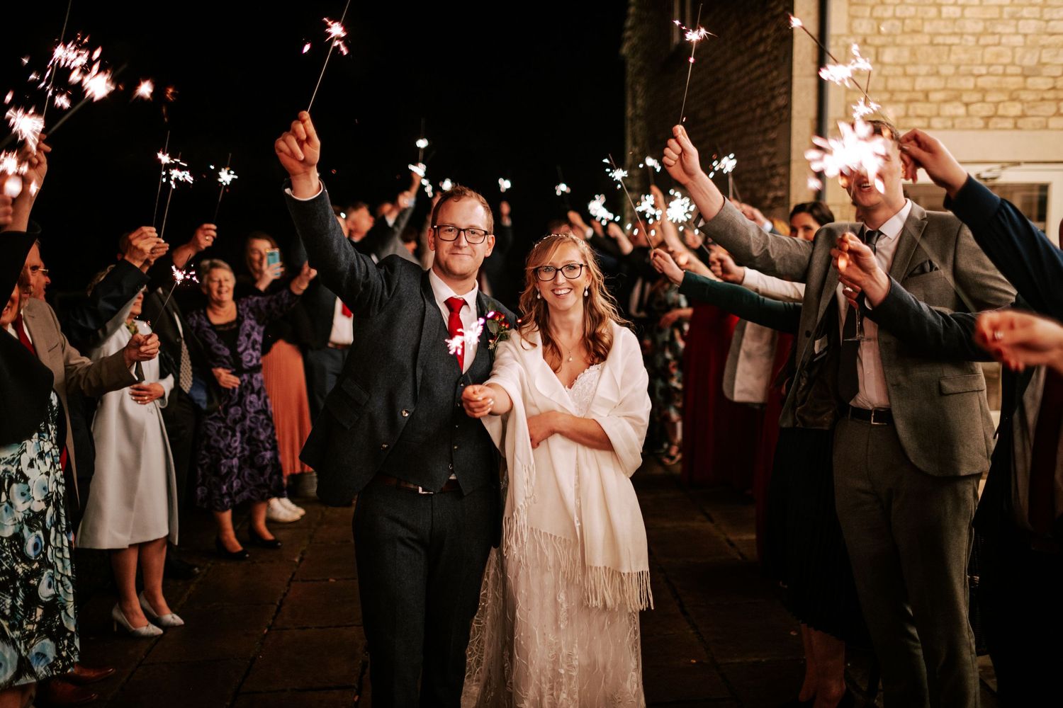 Wedding celebration at night with guests holding sparklers creating a magical exit tunnel.