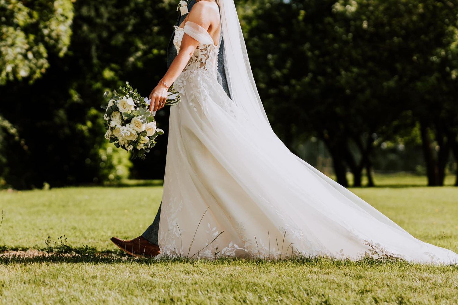 Flowing white wedding dress with lace details and long train photographed in sunlit garden.