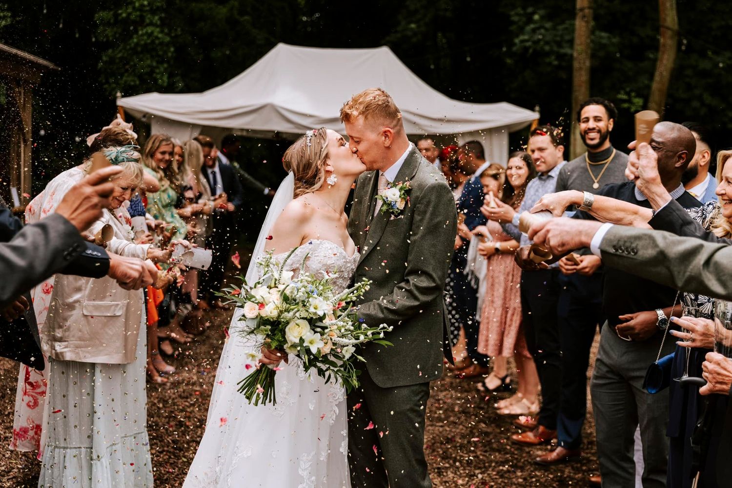 Wedding guests forming a celebratory tunnel with raised arms outdoors.