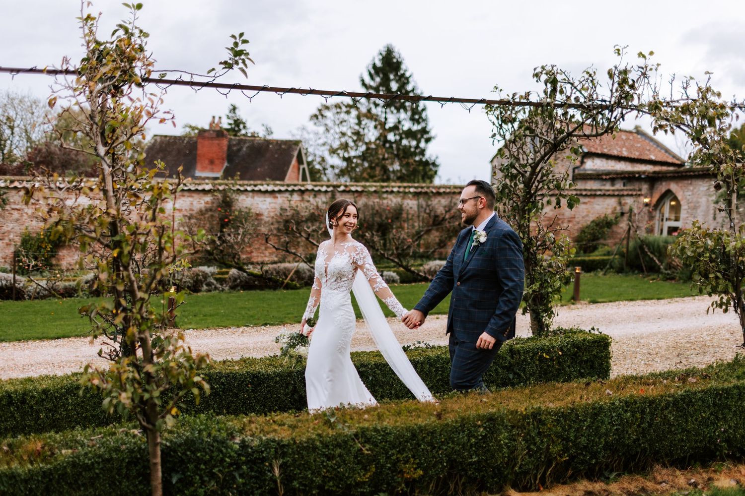 Newlyweds walking and holding hands in a garden pathway surrounded by young trees.