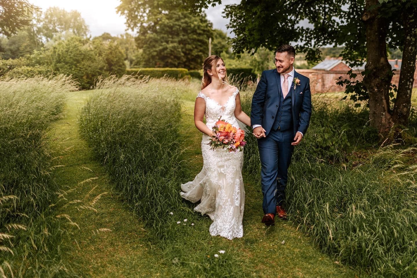 Newlyweds walking through tall grass in garden setting during summer wedding photoshoot.