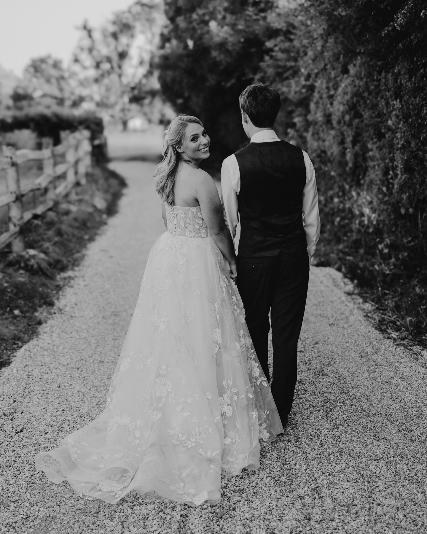 Black and white photo of couple walking down garden path in wedding attire.