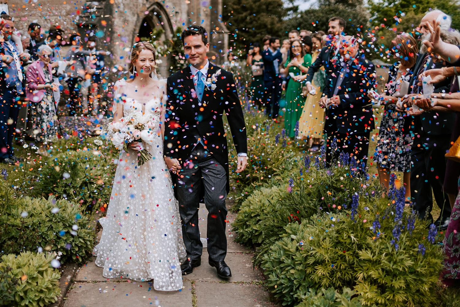 Newlyweds walk through shower of confetti in garden path while guests celebrate.