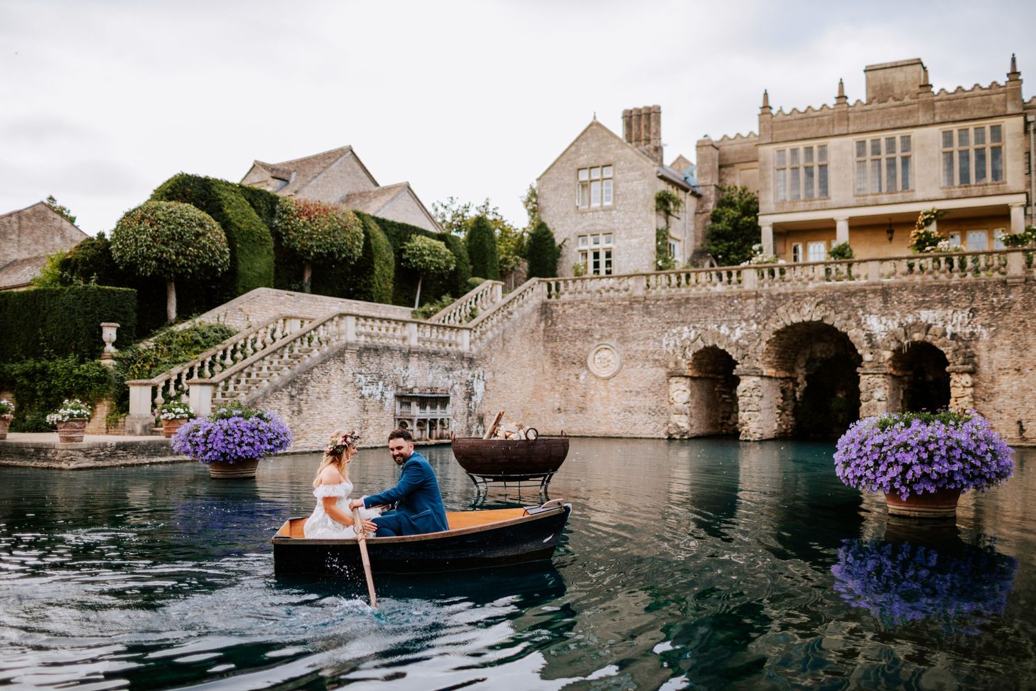 Couple rowing boat on castle moat surrounded by stone architecture and purple flowers.