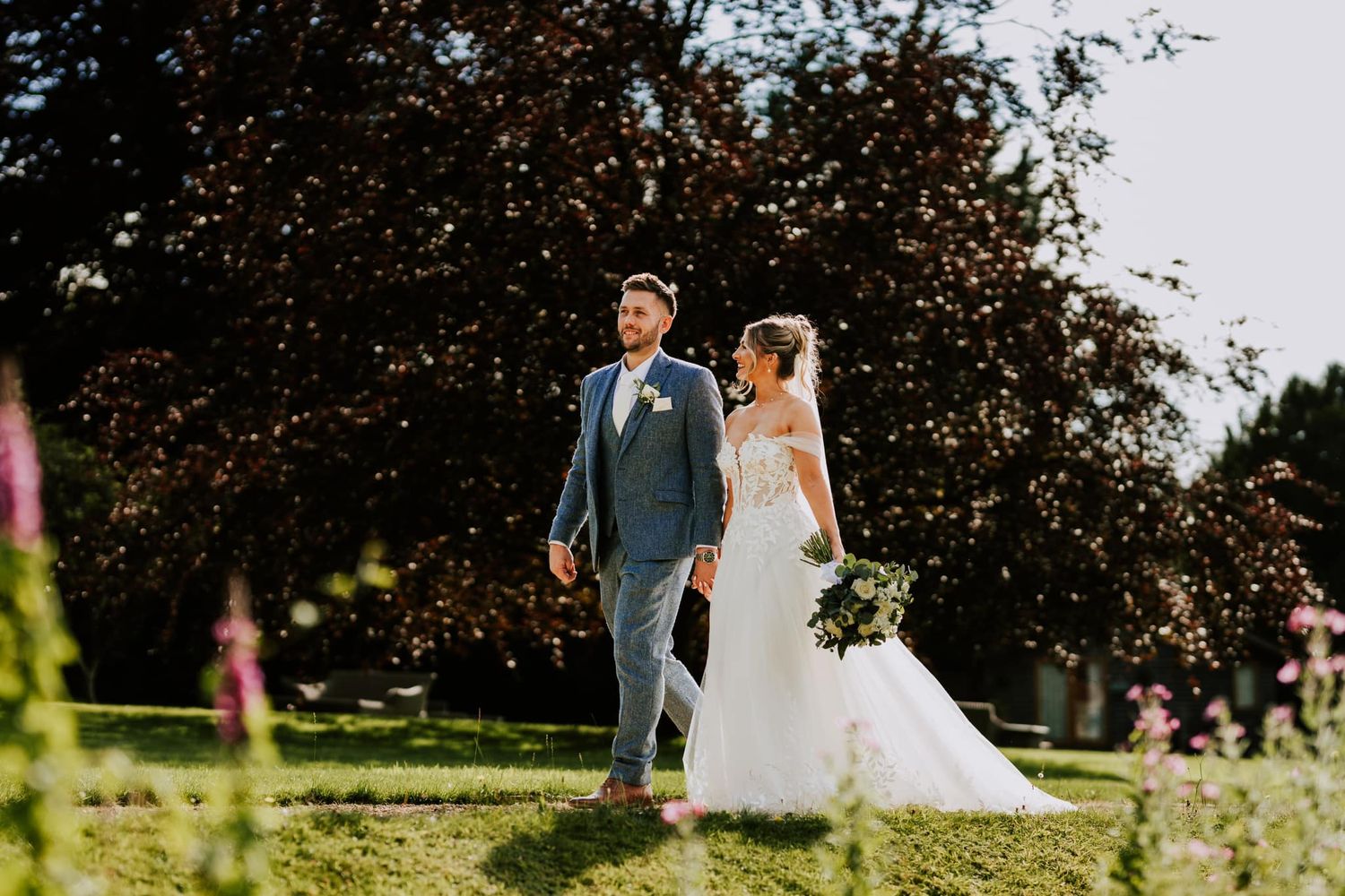 A couple strolls through a sun-drenched garden on their wedding day with flowers in the foreground.