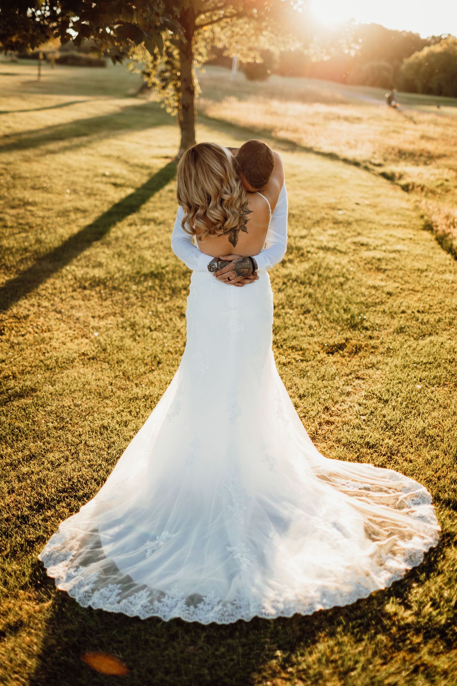 Bride's detailed lace wedding dress train spreads beautifully across sun-drenched grass.