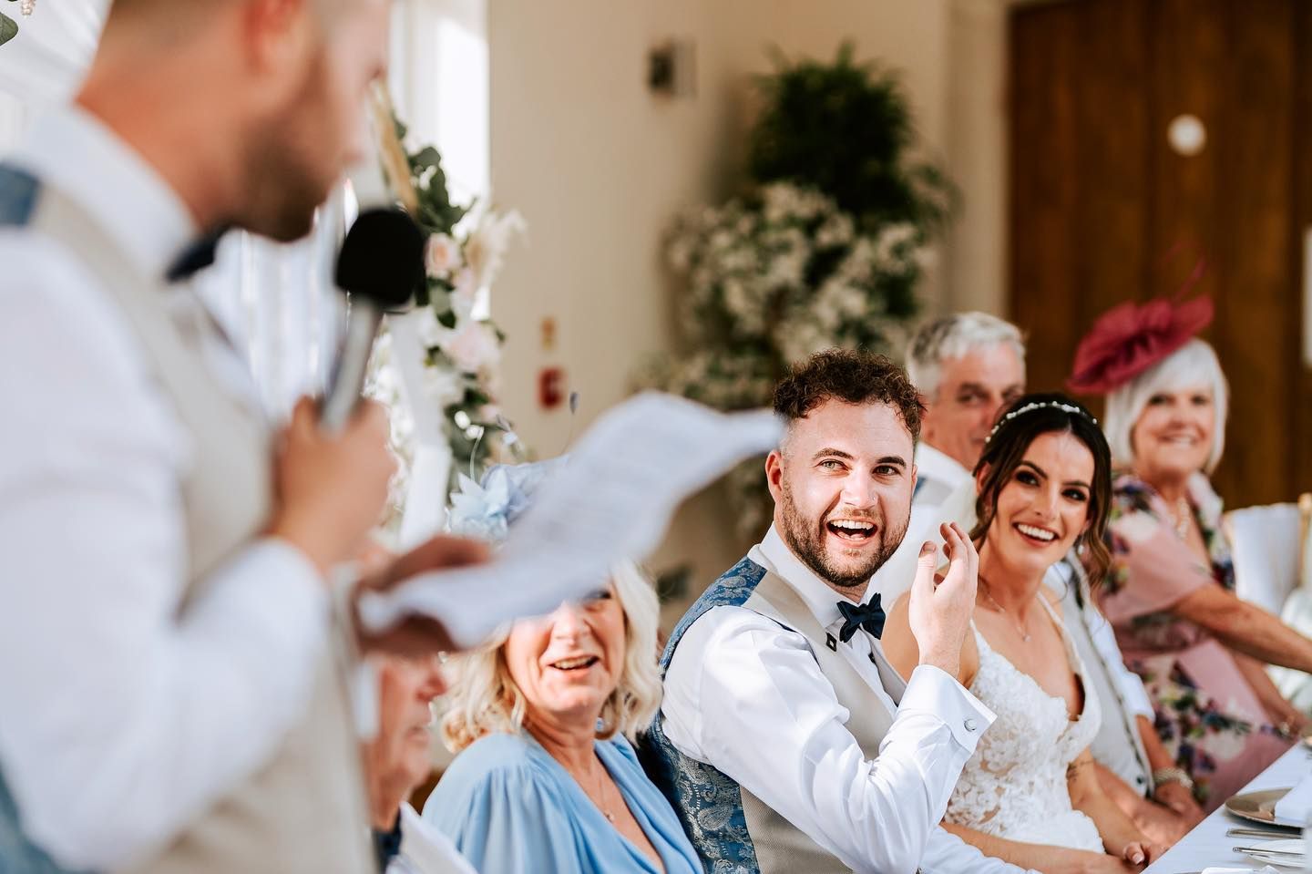 Wedding guests laugh joyfully during reception speeches in an elegantly decorated venue.