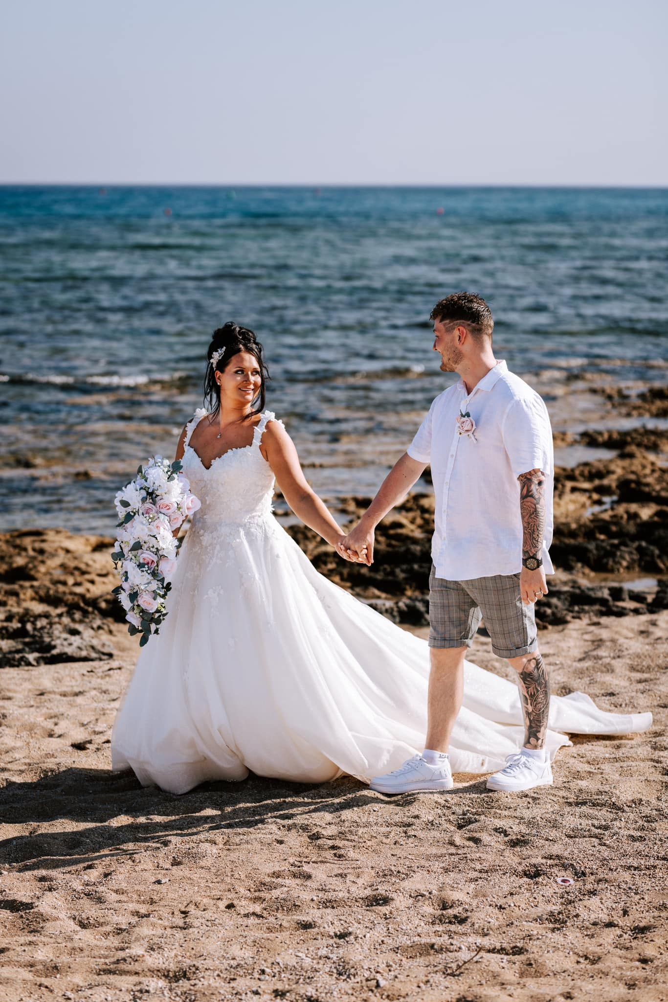 Couple holds hands on beach, bride in flowing white dress against ocean backdrop.