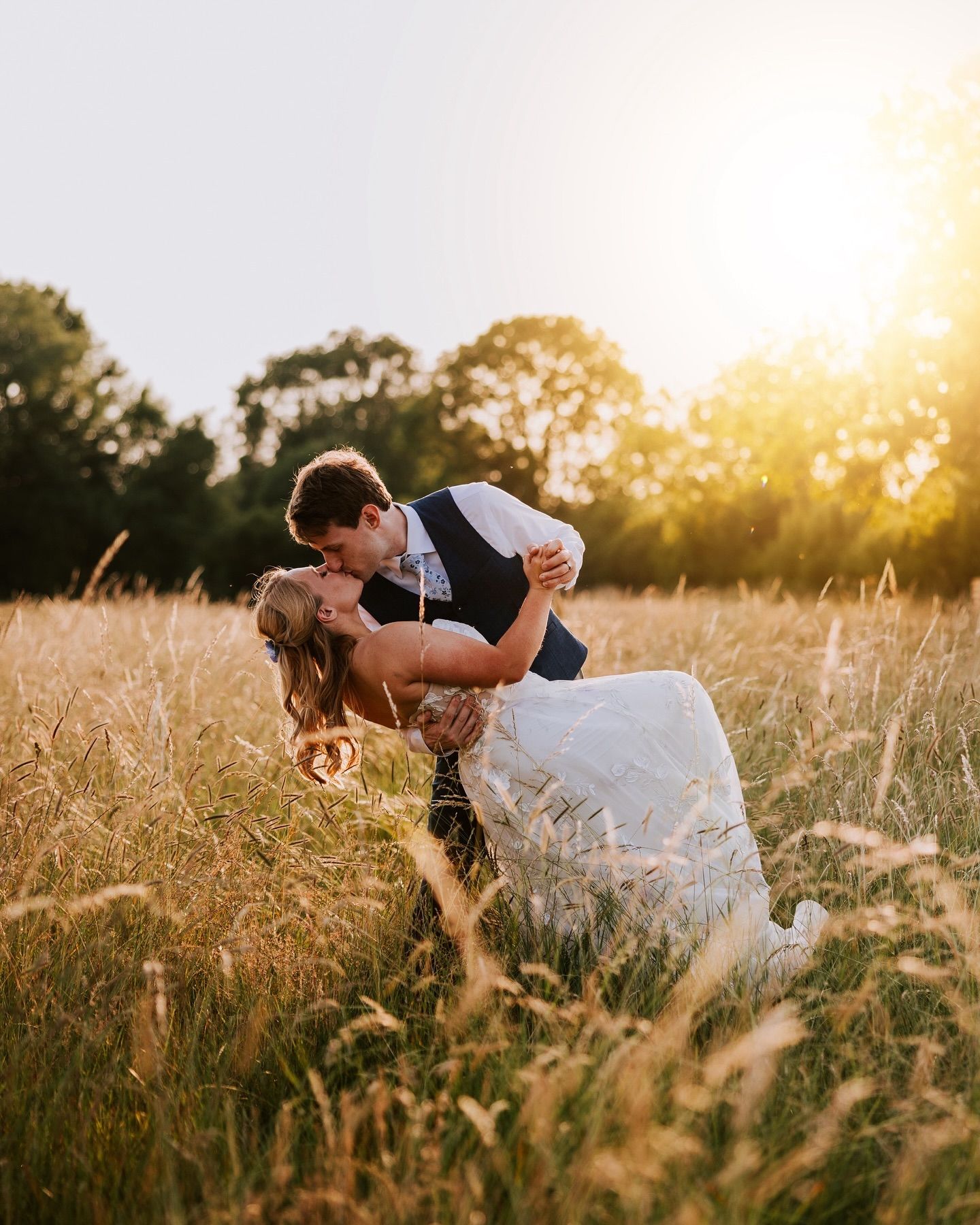 Romantic wedding couple embraces in golden sunset field during dramatic dip pose.