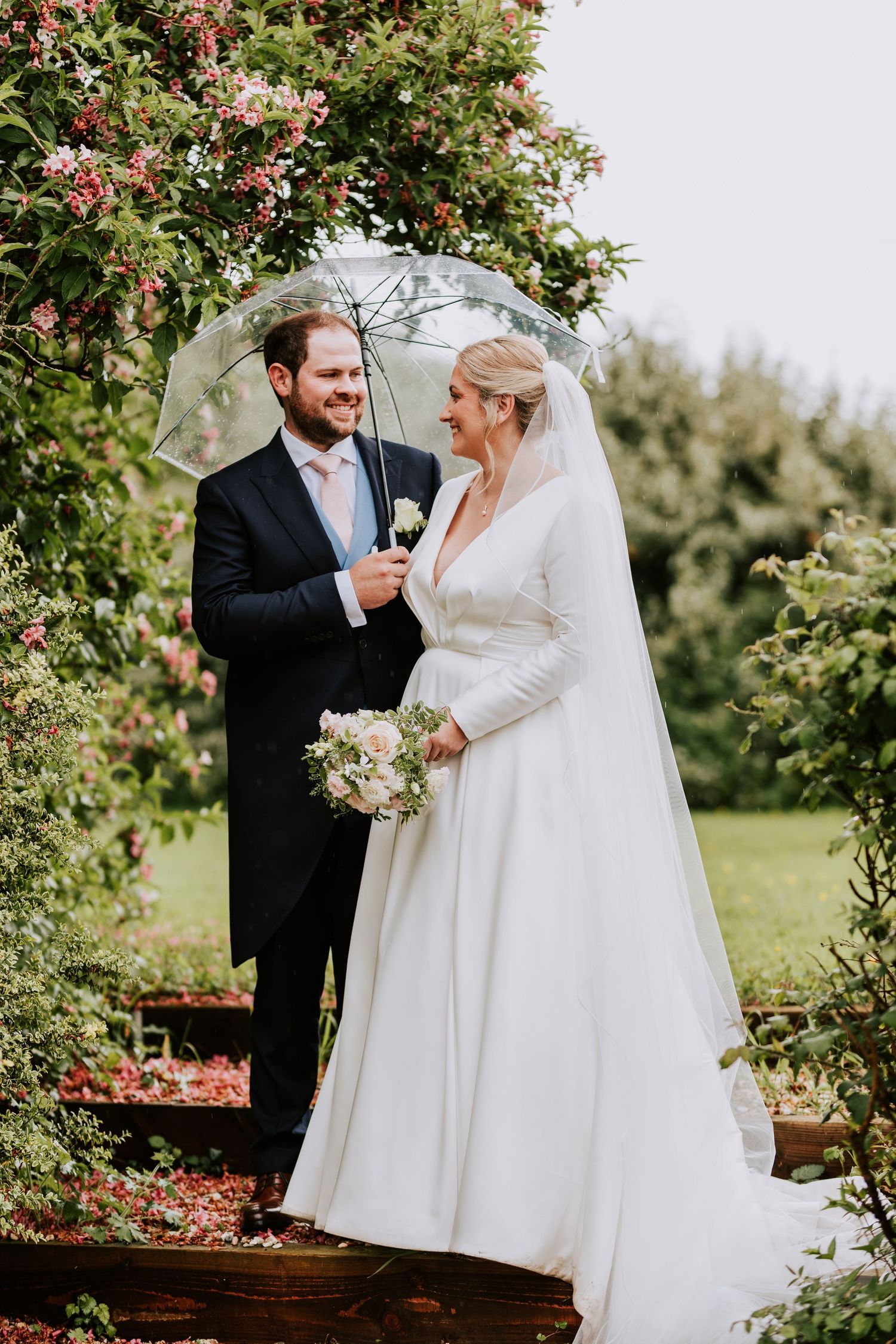 A wedding couple shares a joyful moment together surrounded by blooming red flowers in an outdoor garden setting.