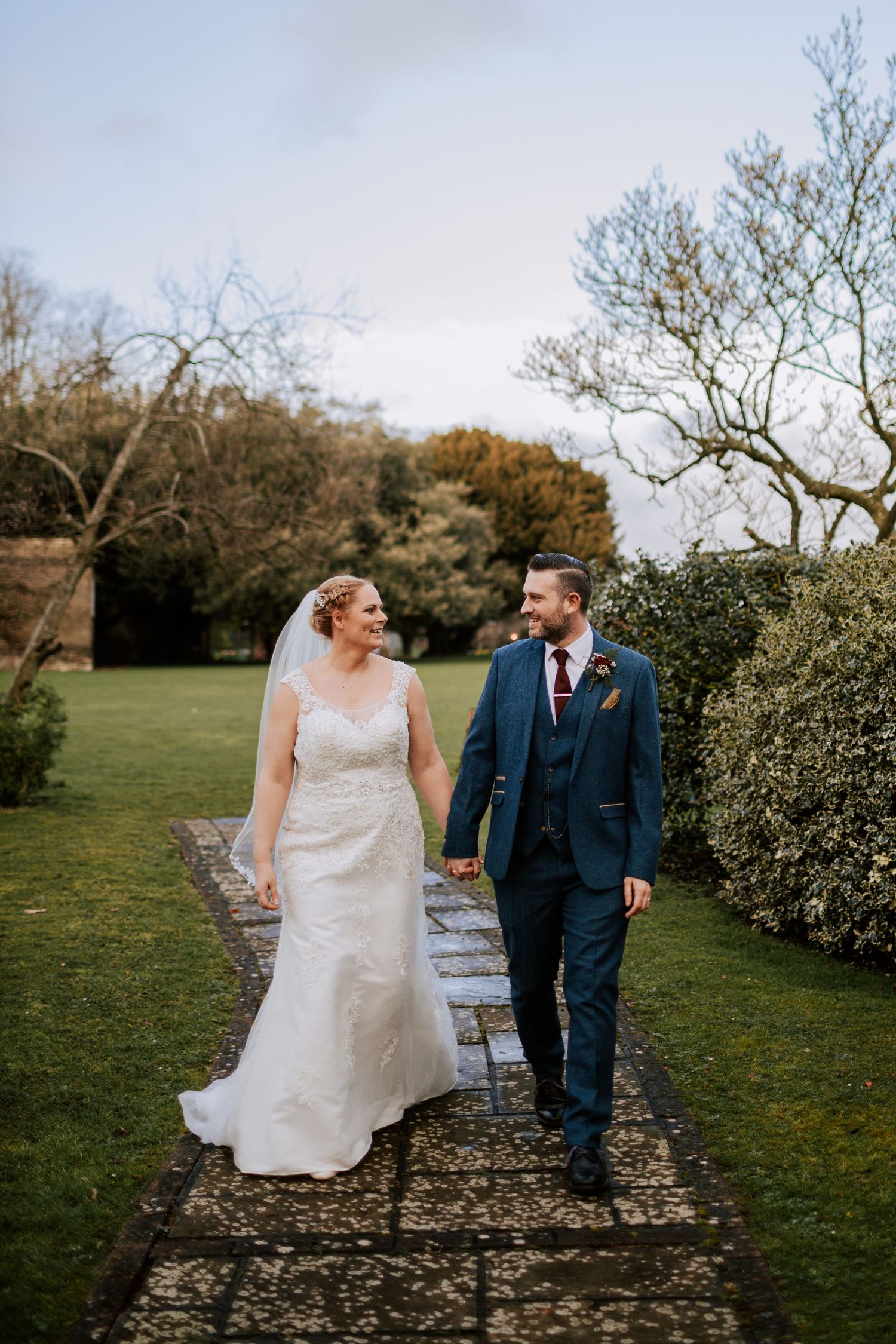 Newlyweds walk hand-in-hand along a garden path surrounded by lush greenery at sunset.