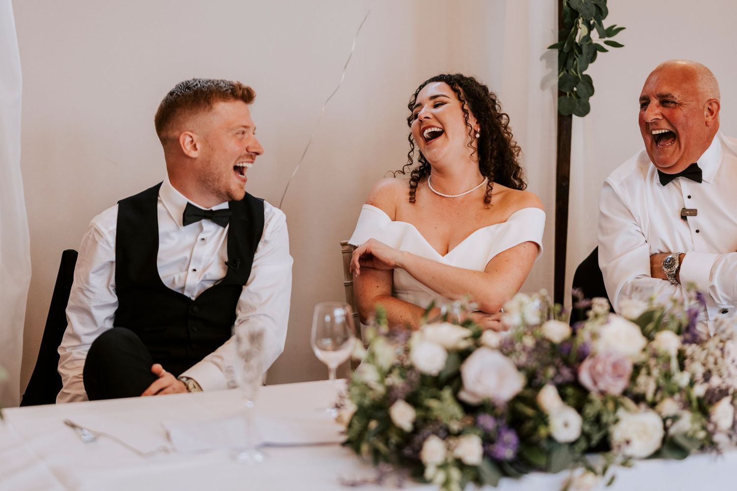 Wedding reception table scene with couple laughing among floral centerpieces.
