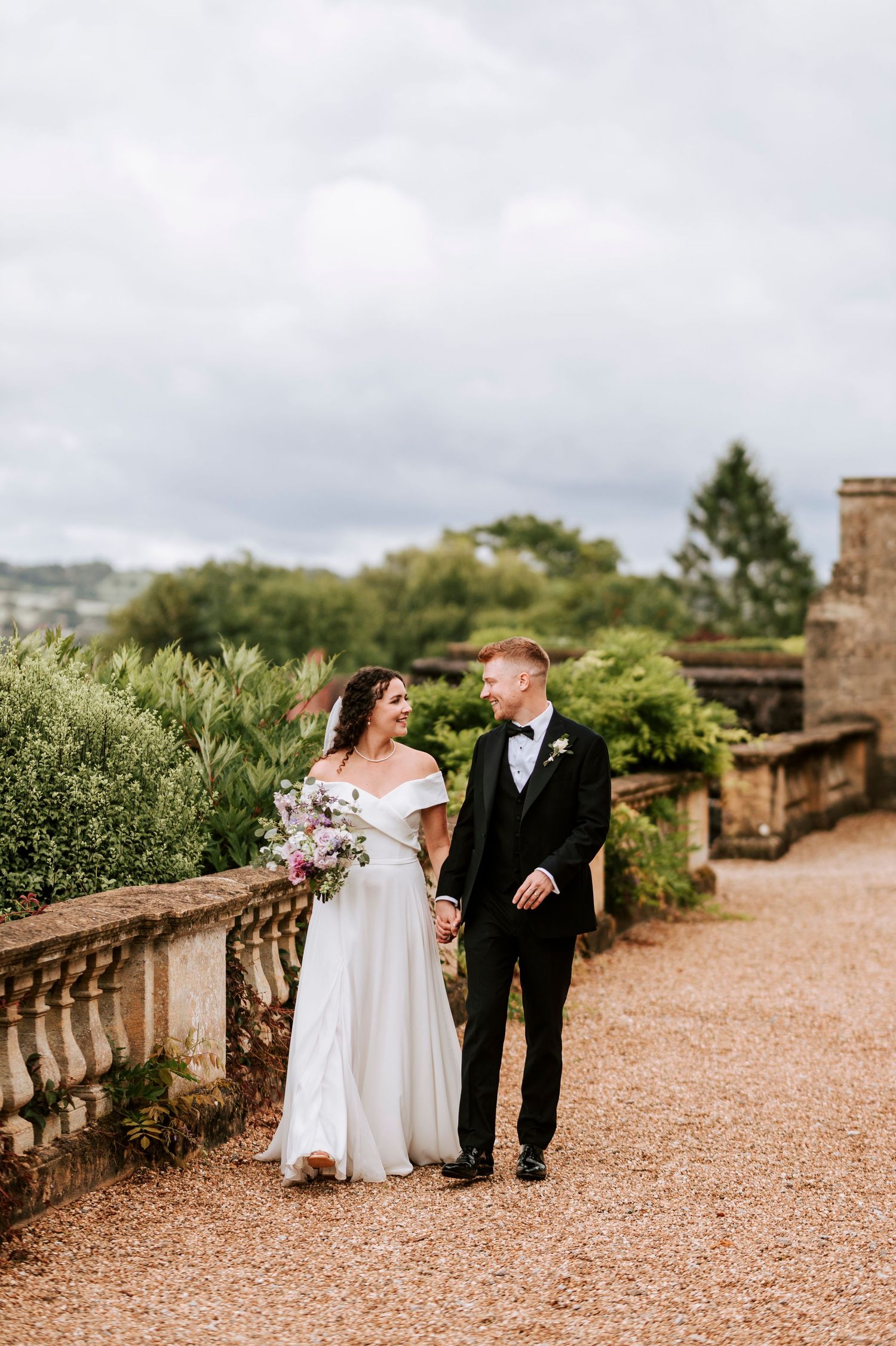 Couple walks along gravel path surrounded by greenery and stone architecture.