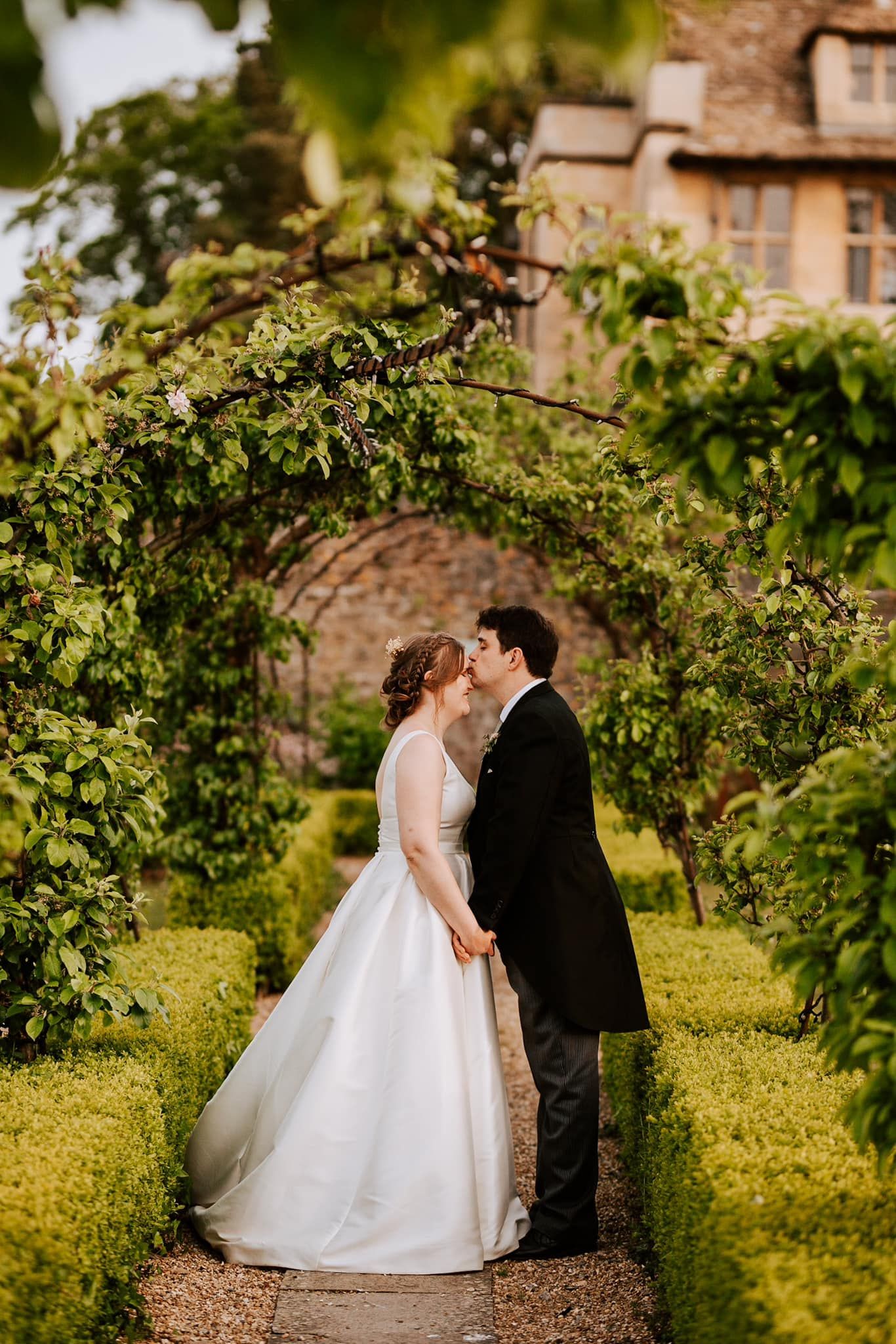 Couple shares intimate moment under garden archway with greenery and stone building backdrop.