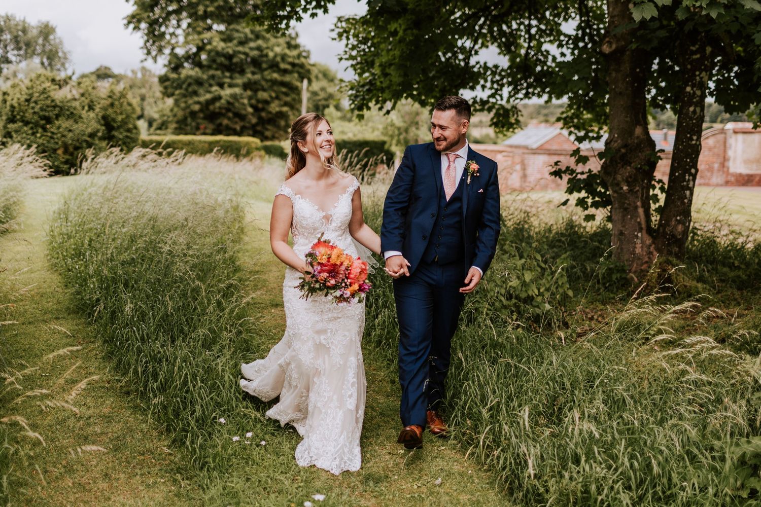 A wedding couple walks through tall grass on a sunny summer day.