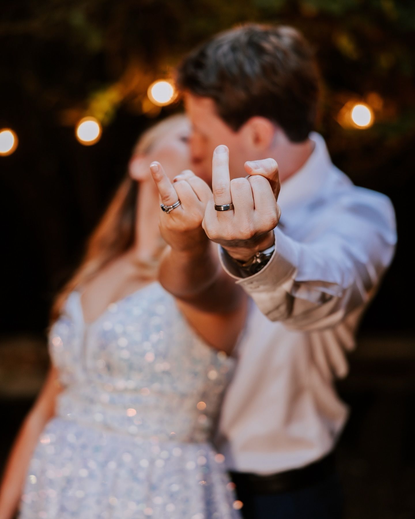 Wedding couple shows off their rings in playful pose with string lights in background.