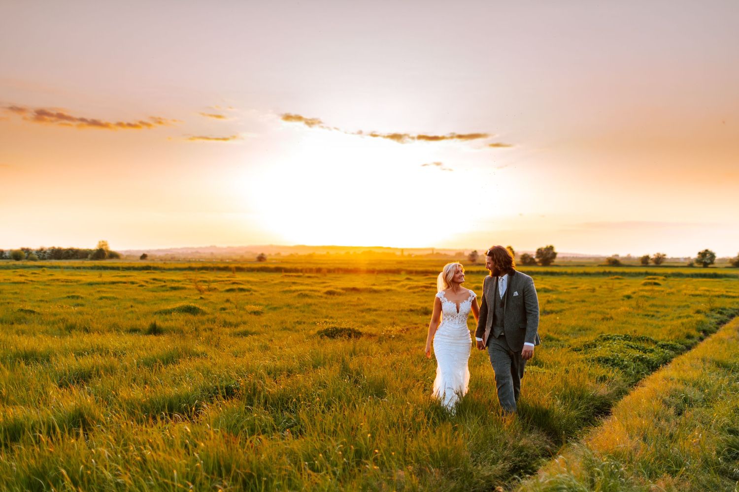 A couple walks through a golden wheat field at sunset with warm glowing light in the background.