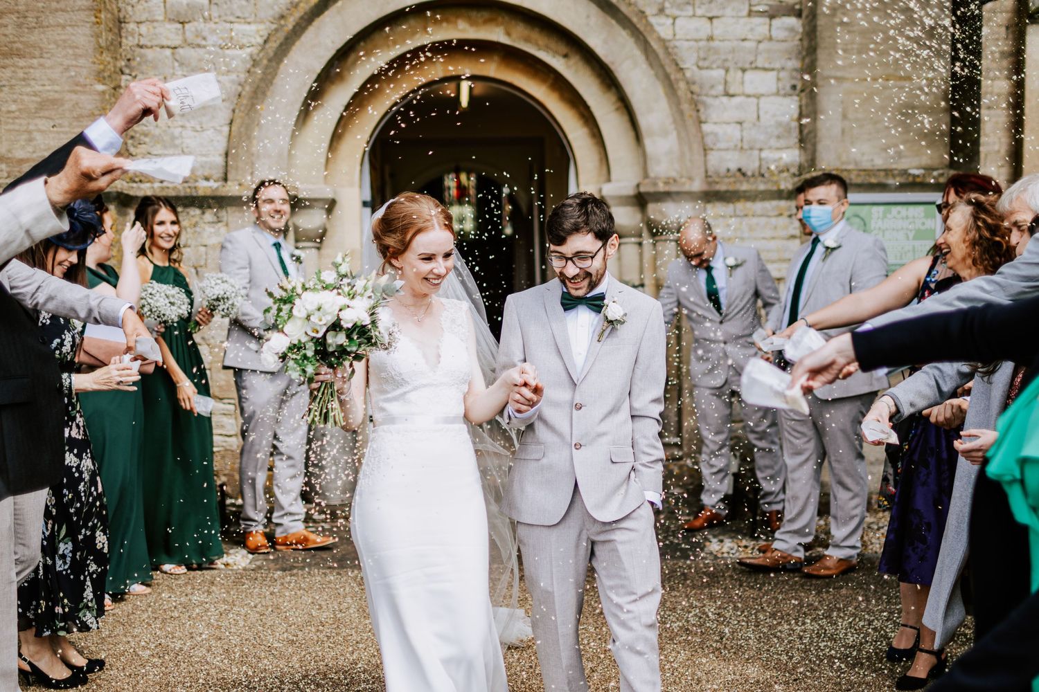 Wedding party celebrates with confetti throw outside historic stone church entrance.