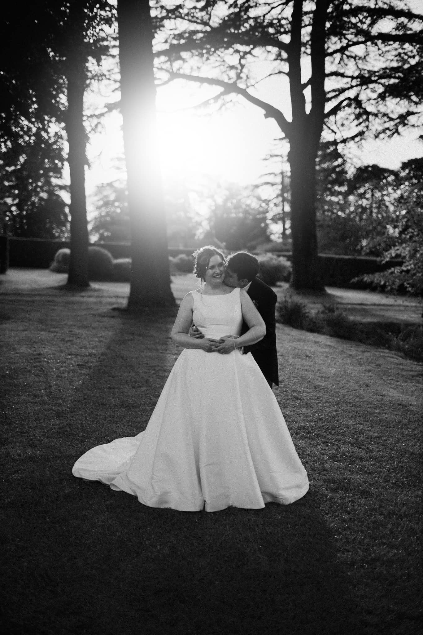 Silhouetted couple embraces during sunset among tall trees in garden setting.