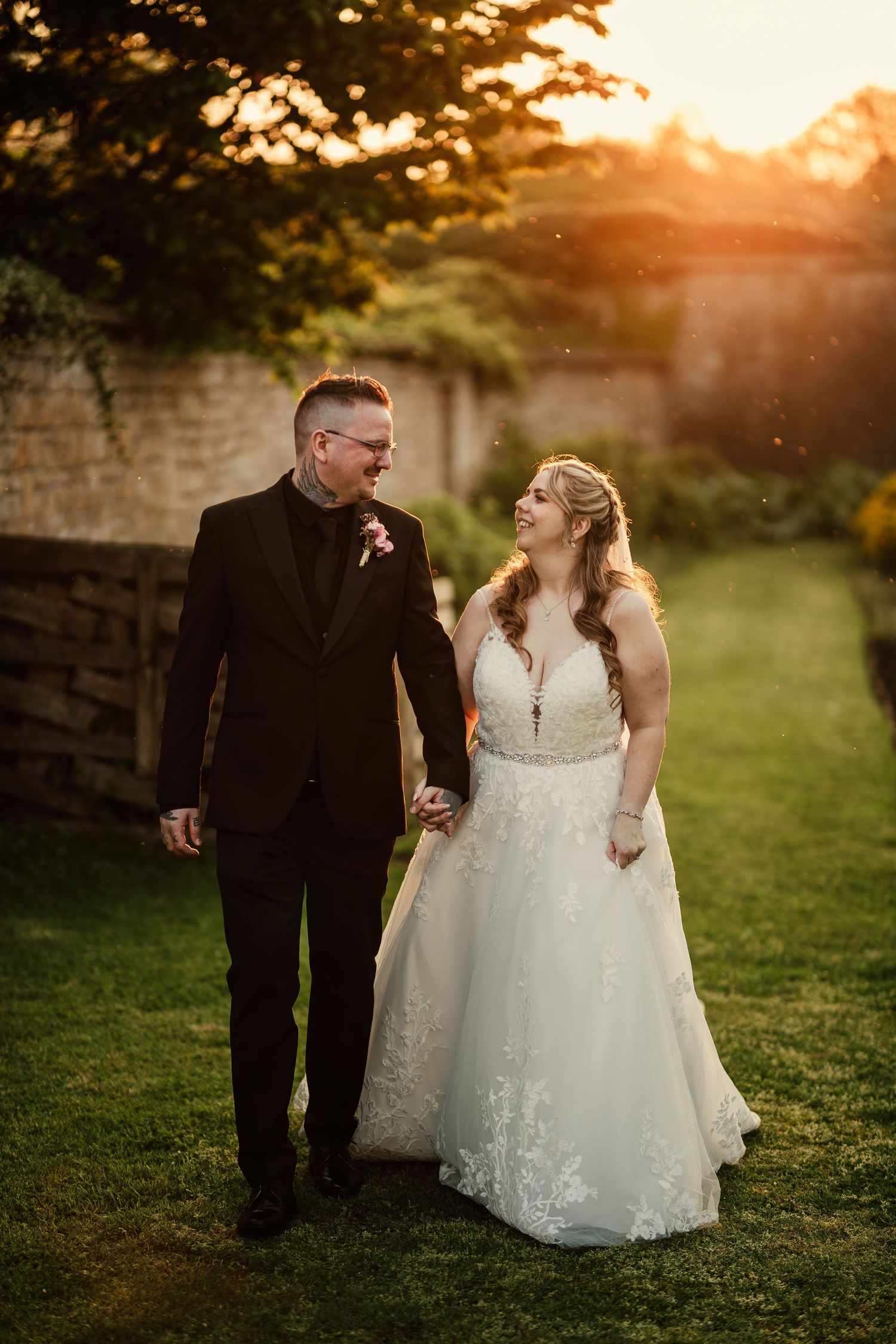 A couple in wedding attire walks together on grass at sunset near a stone wall.