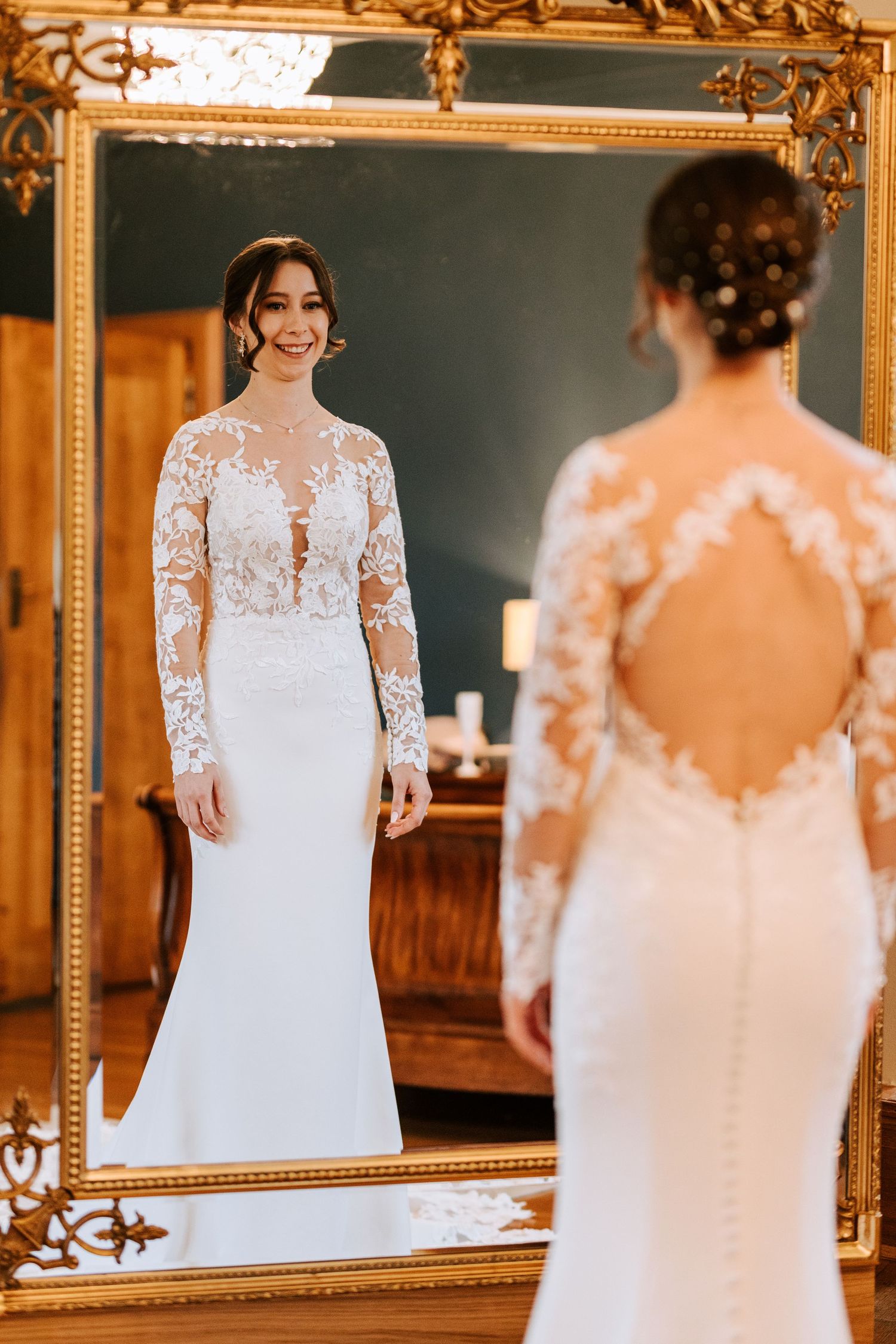 Elegant bride in lace long-sleeved wedding gown with open back detail admiring herself in ornate gold mirror.
