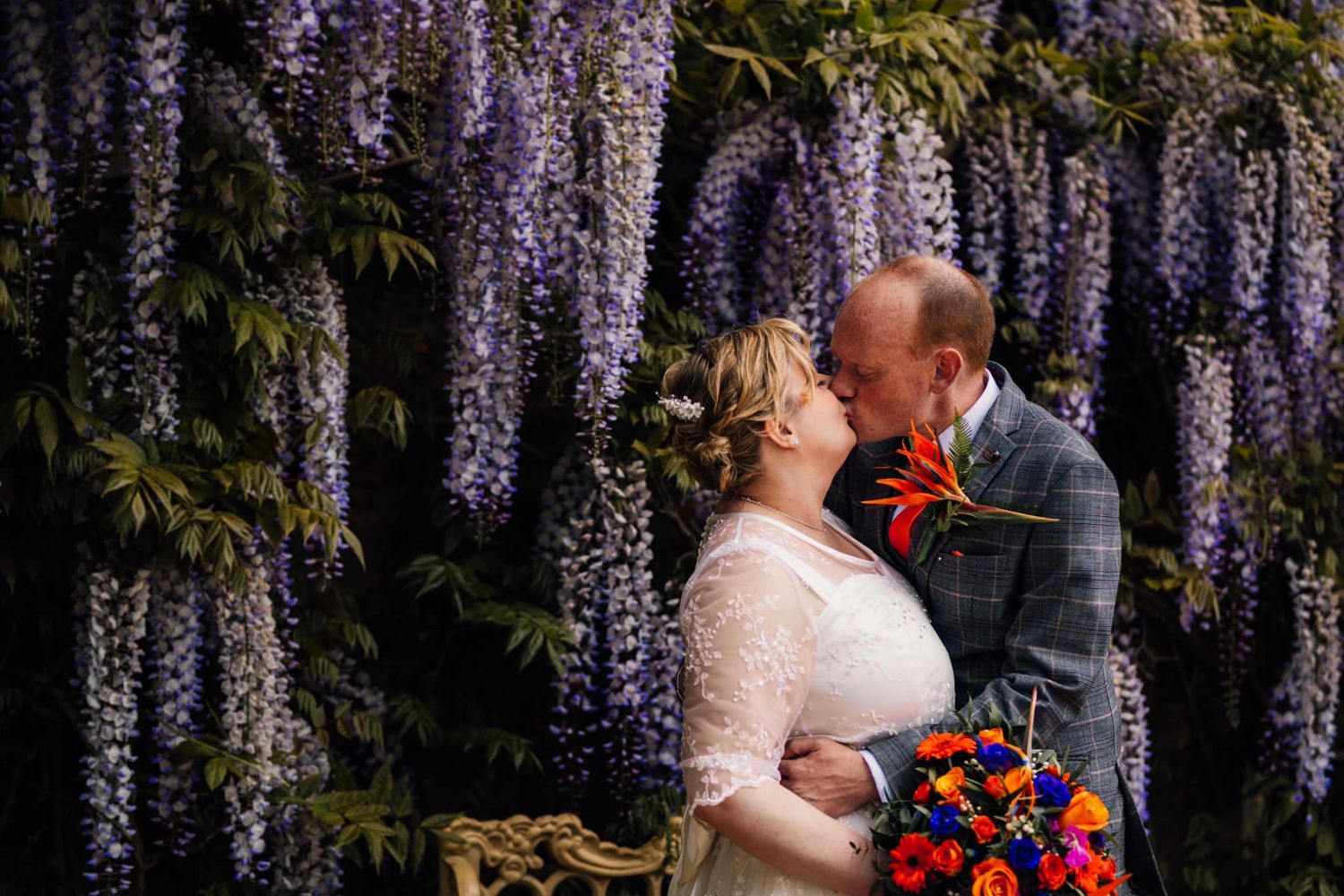 Couple embraces in front of blooming purple wisteria with colorful bouquet.
