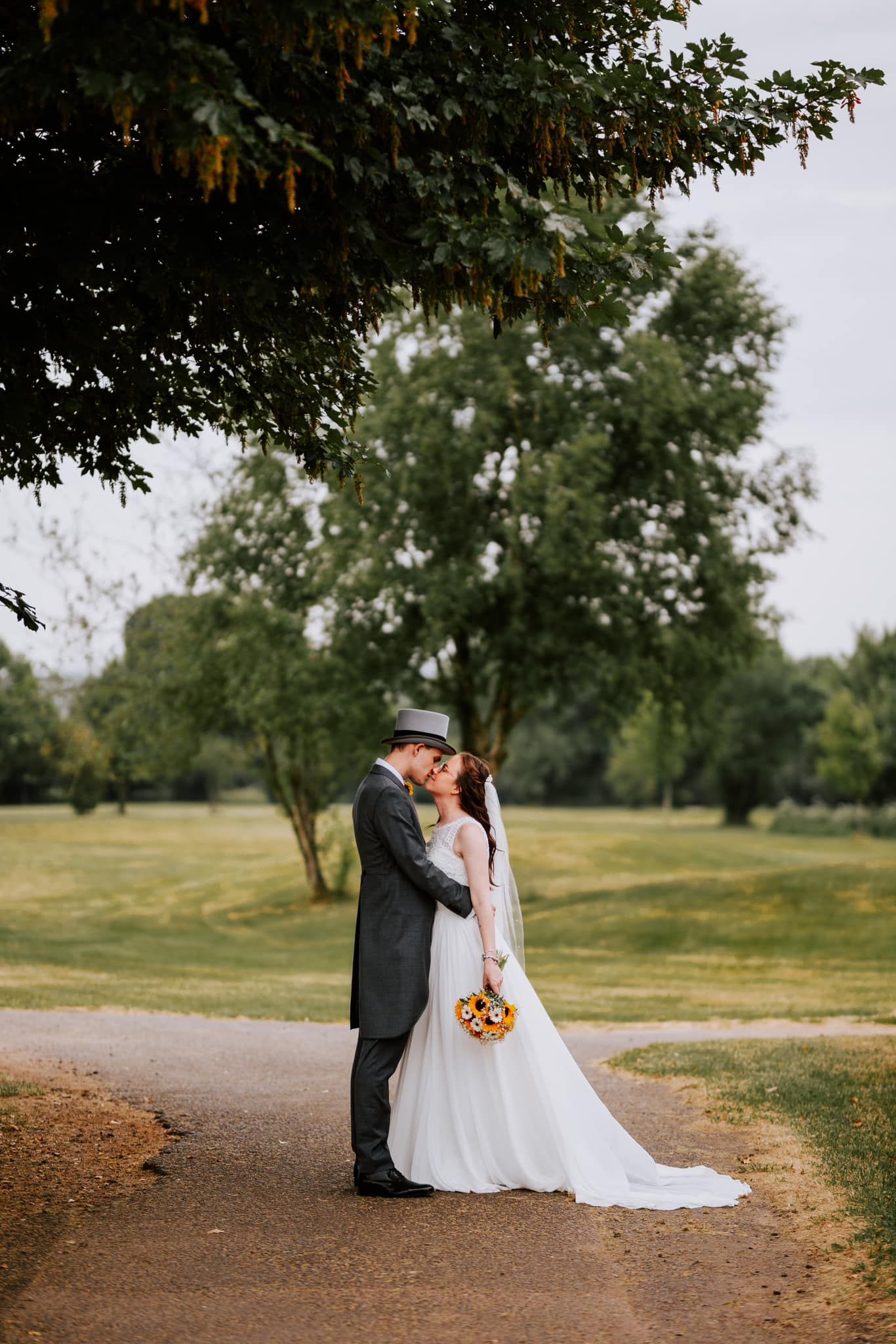 Couple shares romantic moment on dirt path under trees while holding sunflower bouquet.