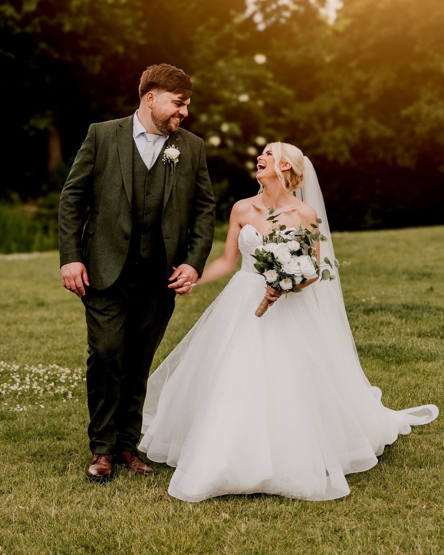 Candid wedding photo of couple laughing together on grassy lawn at sunset.