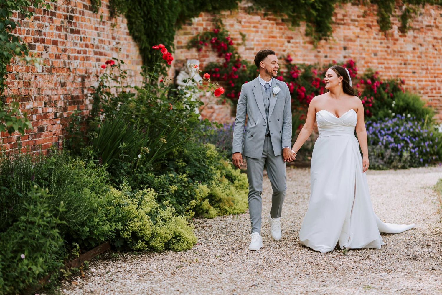 Couple walks hand in hand through garden with brick wall and colorful flowers.