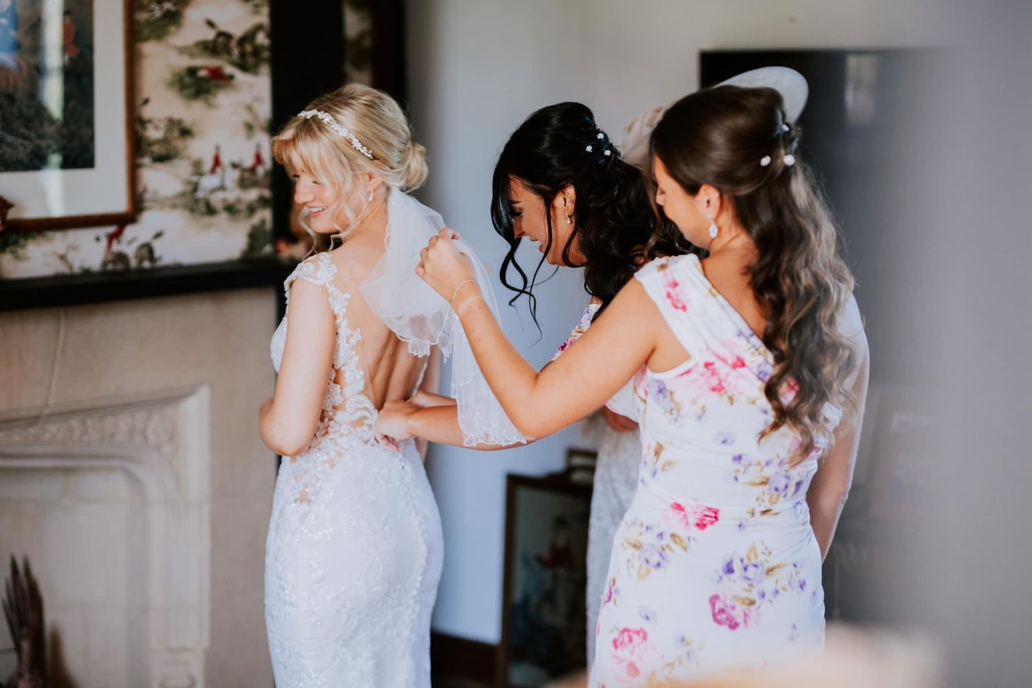 Bridesmaids in floral dresses help zip up a wedding gown during pre-ceremony preparations.