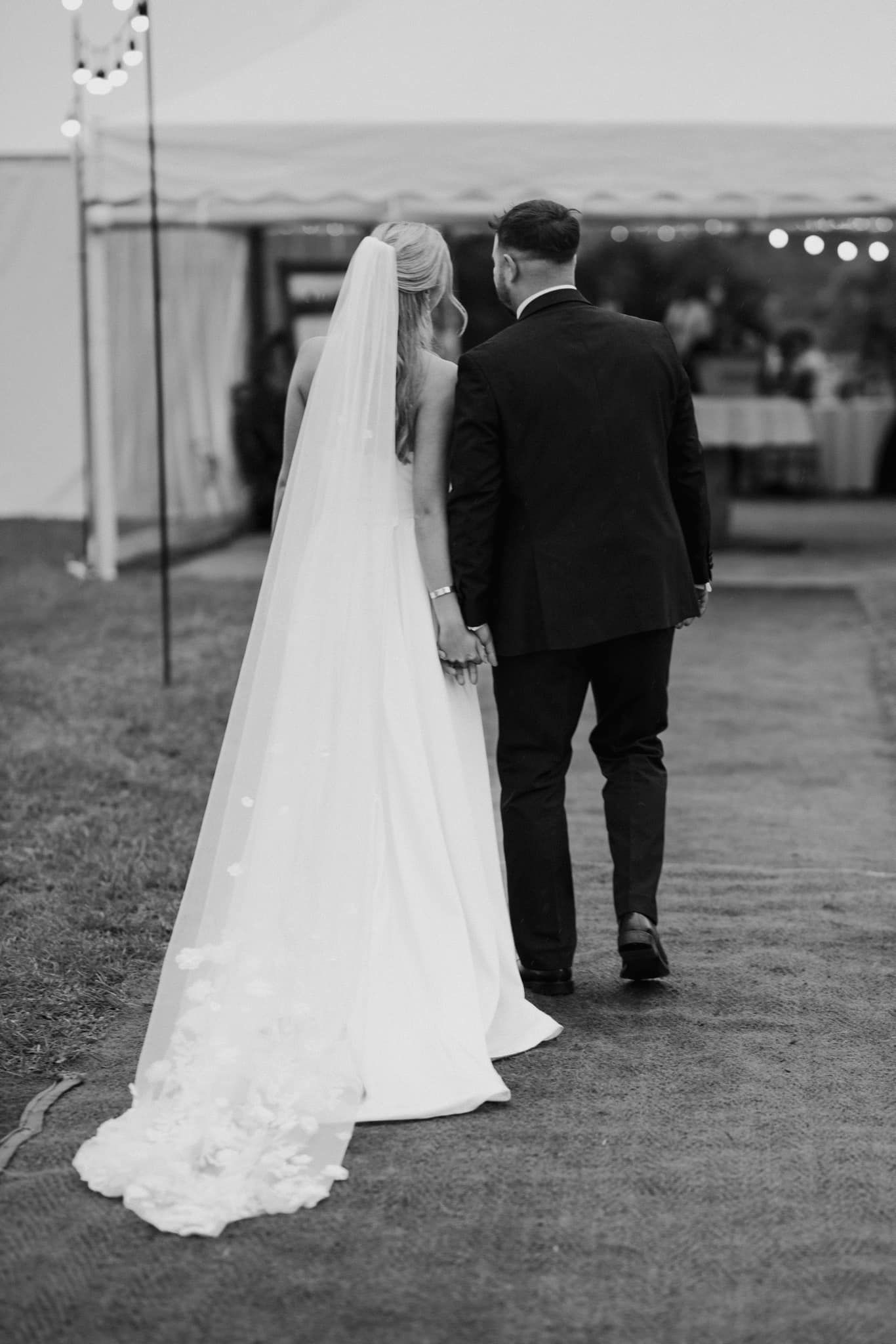 A black and white photo showing a bride in a flowing veil and groom walking away from the camera at an outdoor venue.