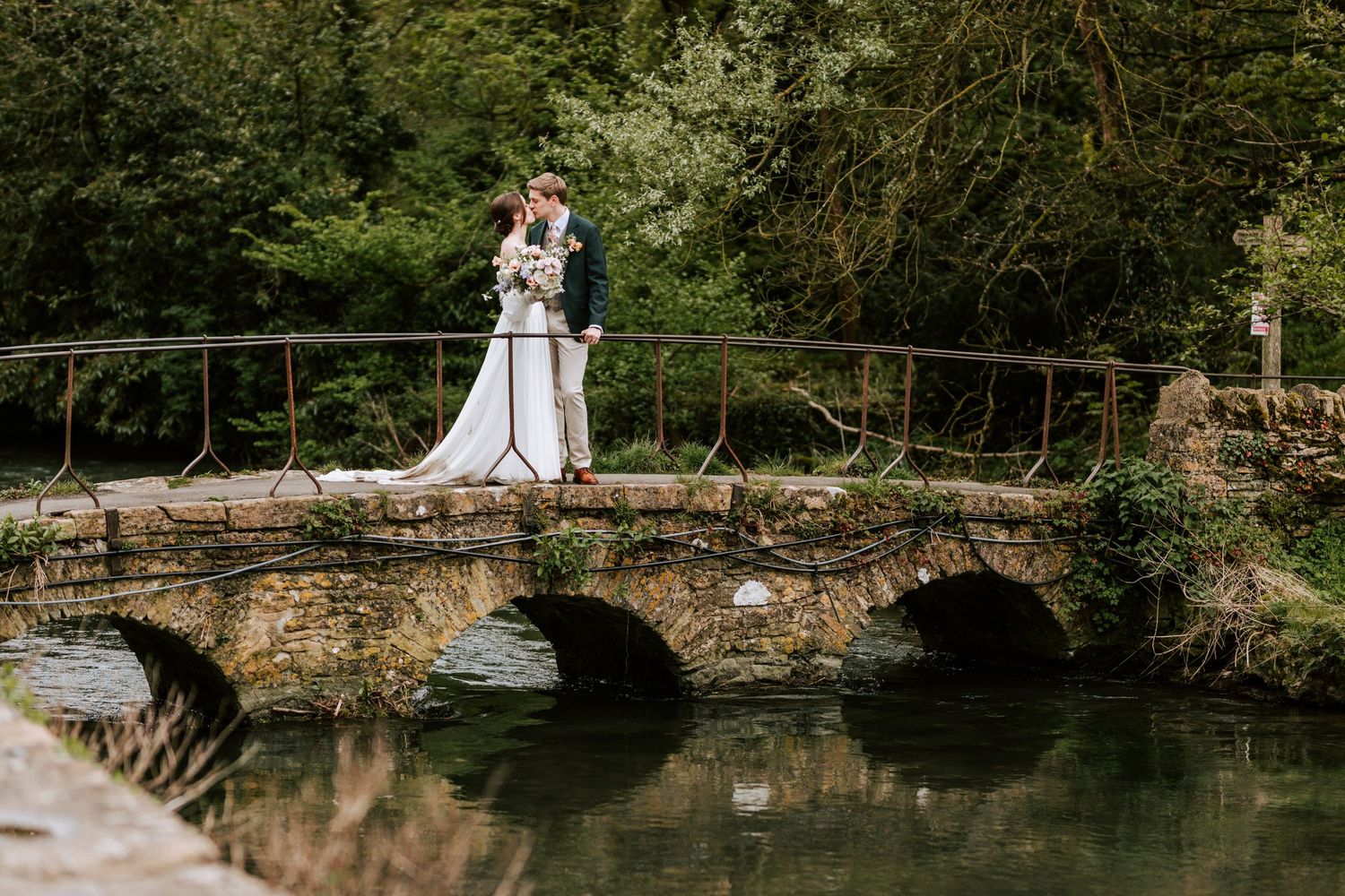 Wedding couple standing on rustic stone bridge over water in garden setting.