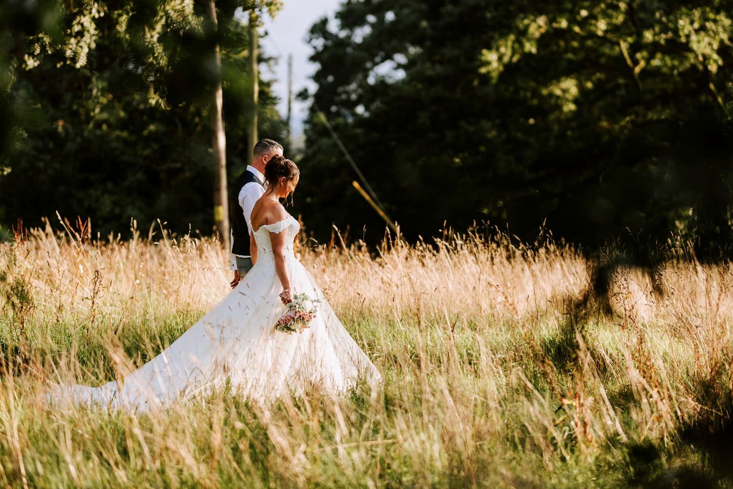 Wedding couple walking through tall grass field at sunset.