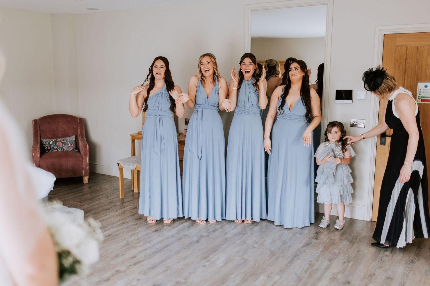 Bridesmaids in flowing light blue dresses react with excitement in a bright indoor setting.