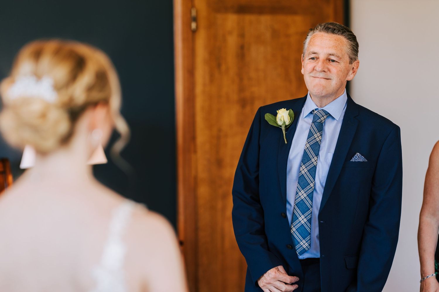 Man in navy suit with plaid tie stands in elegant indoor setting.