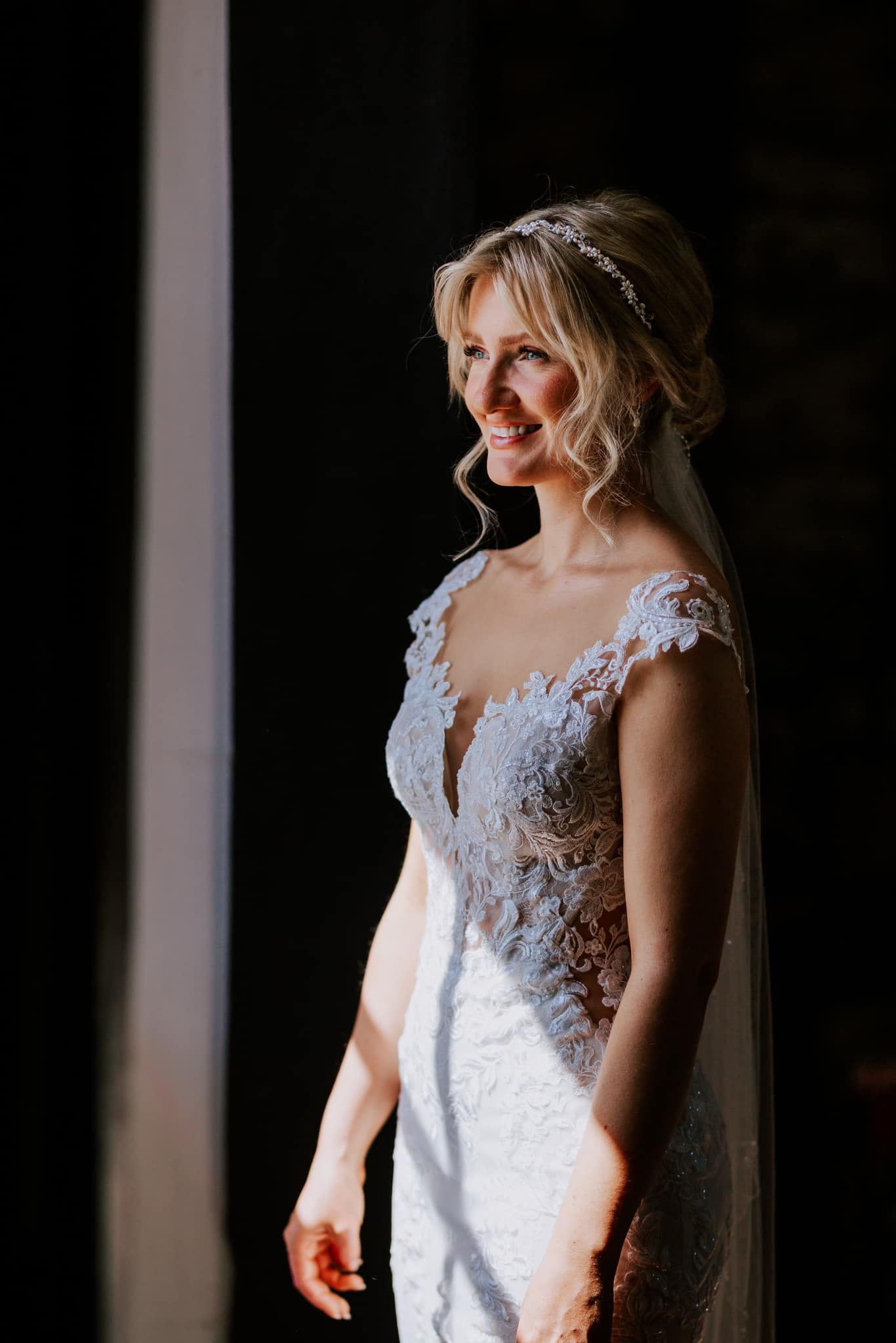 Portrait of bride in lace wedding gown standing by window in natural light.