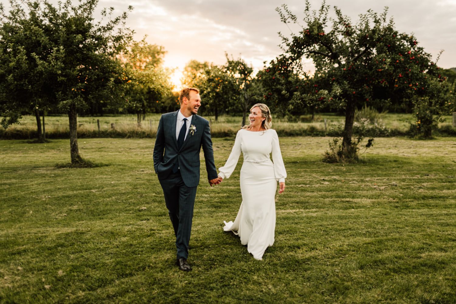 Bride and groom walking hand in hand through green meadow at sunset.