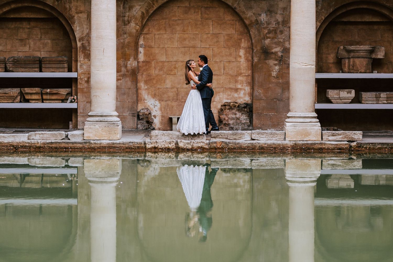 Elegant wedding portraits taken under historic stone archways beside reflecting pool.