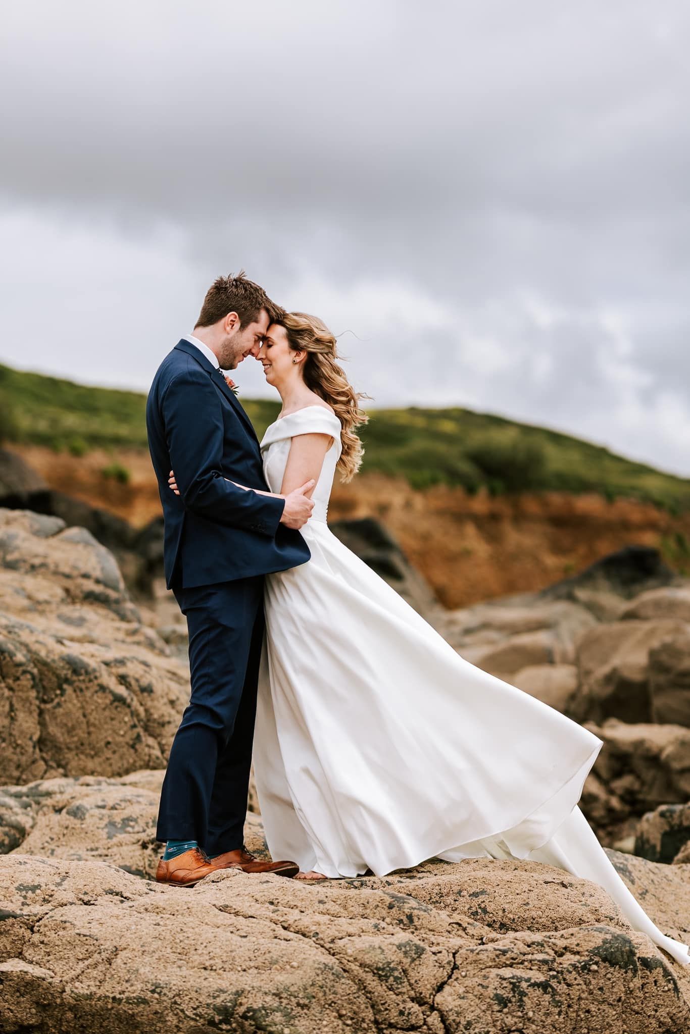 Couple embraces on rocky coastal cliffs with dramatic cloudy sky.