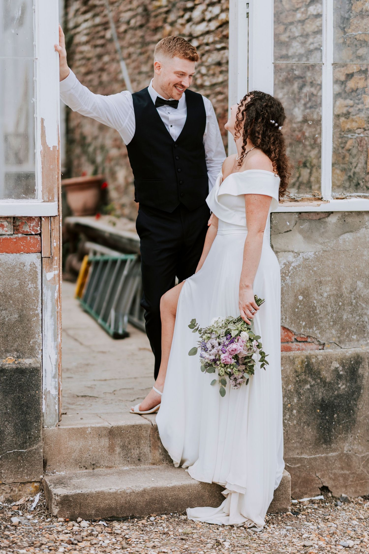 Wedding couple poses near stone wall and glass windows in vintage setting.