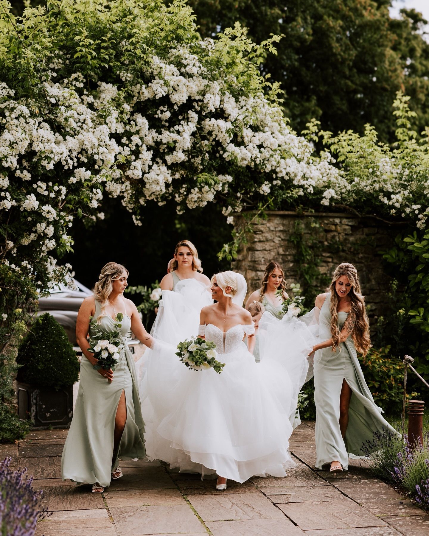 Bridesmaids in sage green dresses help bride with dress under flowering arch in garden.